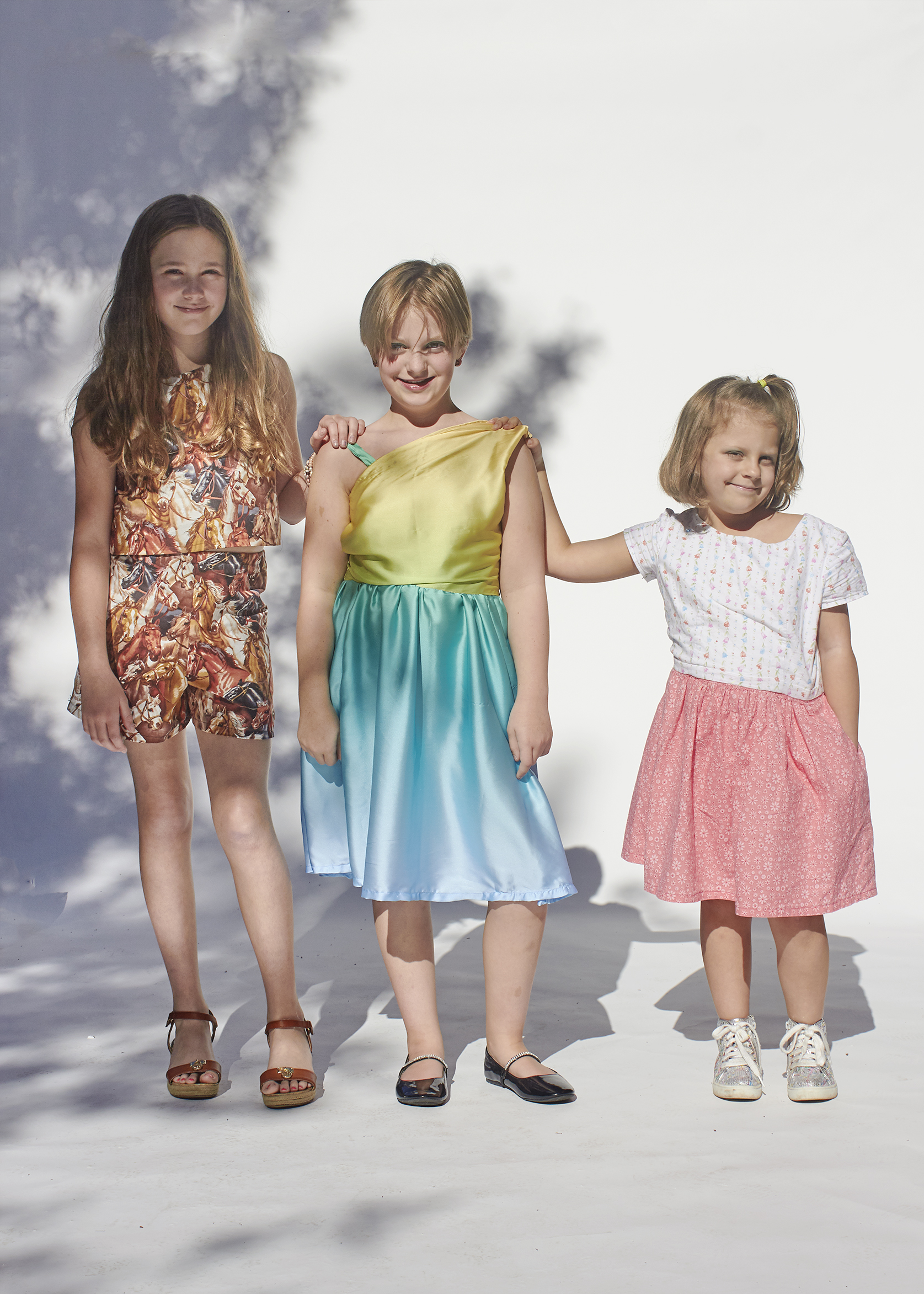 Three girls standing in handmade clothes. The left outfits is a top and shorts made with a horse print. The middle dress is a sunset dress, and the last outfit is a floral top and skirt