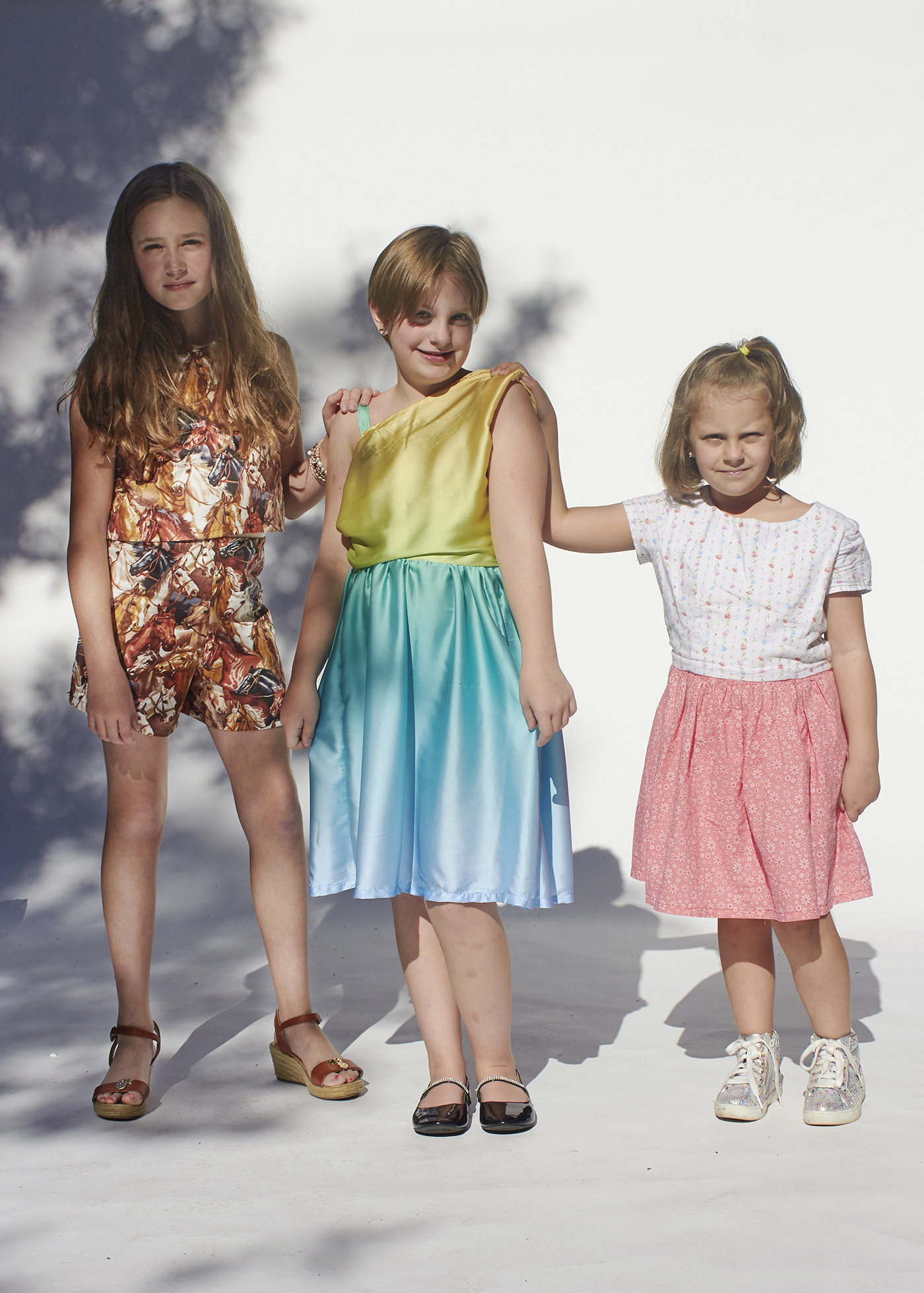 Three girls standing in handmade clothes. The left outfits is a top and shorts made with a horse print. The middle dress is a sunset dress, and the last outfit is a floral top and skirt. Girls have there hands on the shoulder of the middle girl