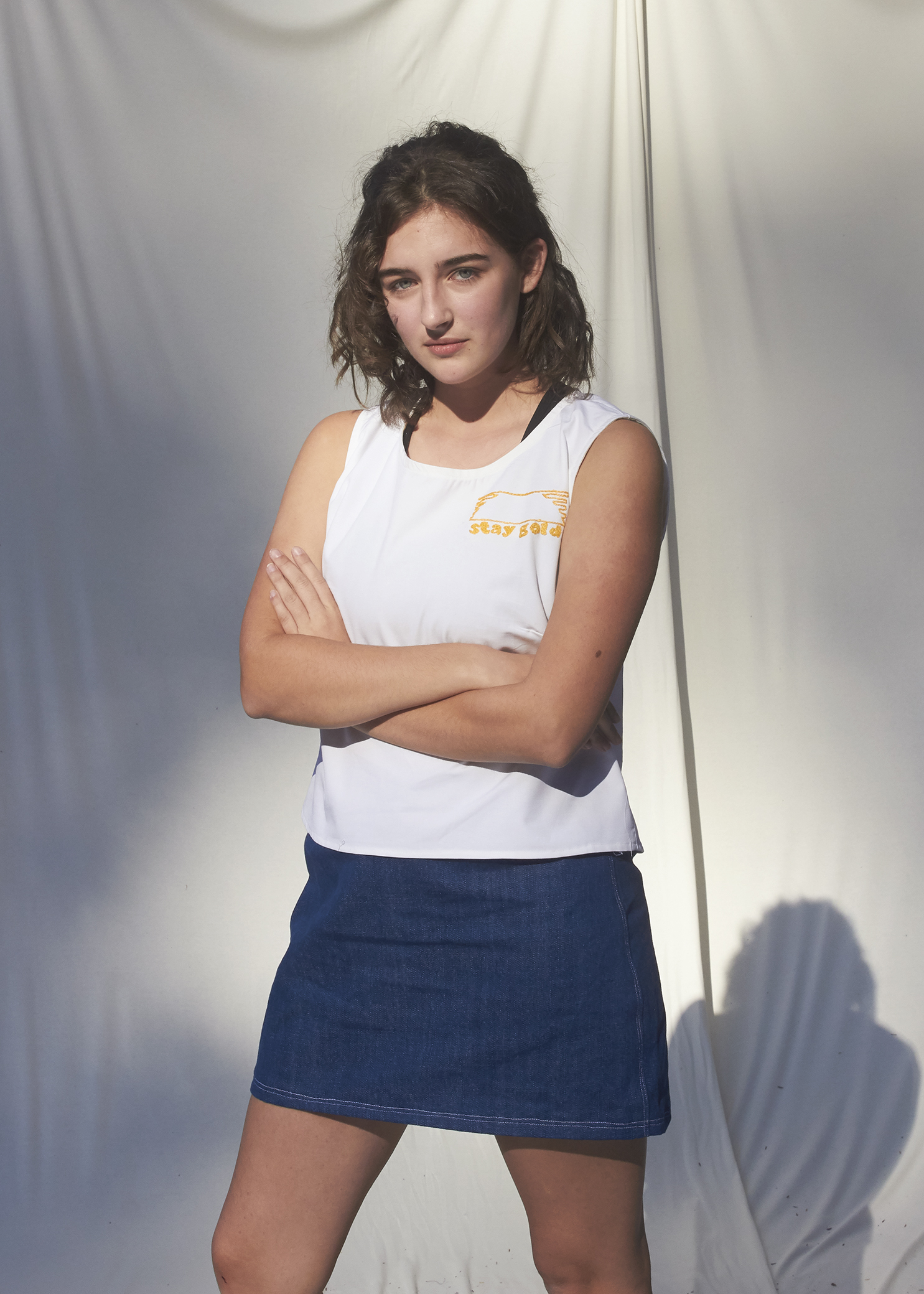 Girl standing in white top with gold embroidery that reads 'Stay Gold' and wears a denim skirt.