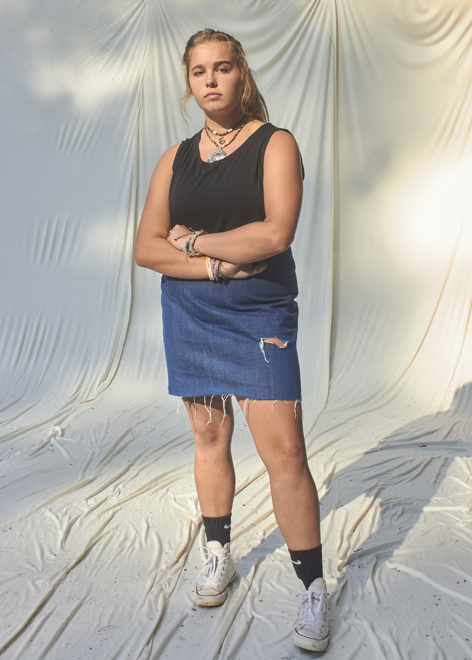 Teenage Girl standing with her arms crossed and is wearing a black top and a denim distressed skirt.