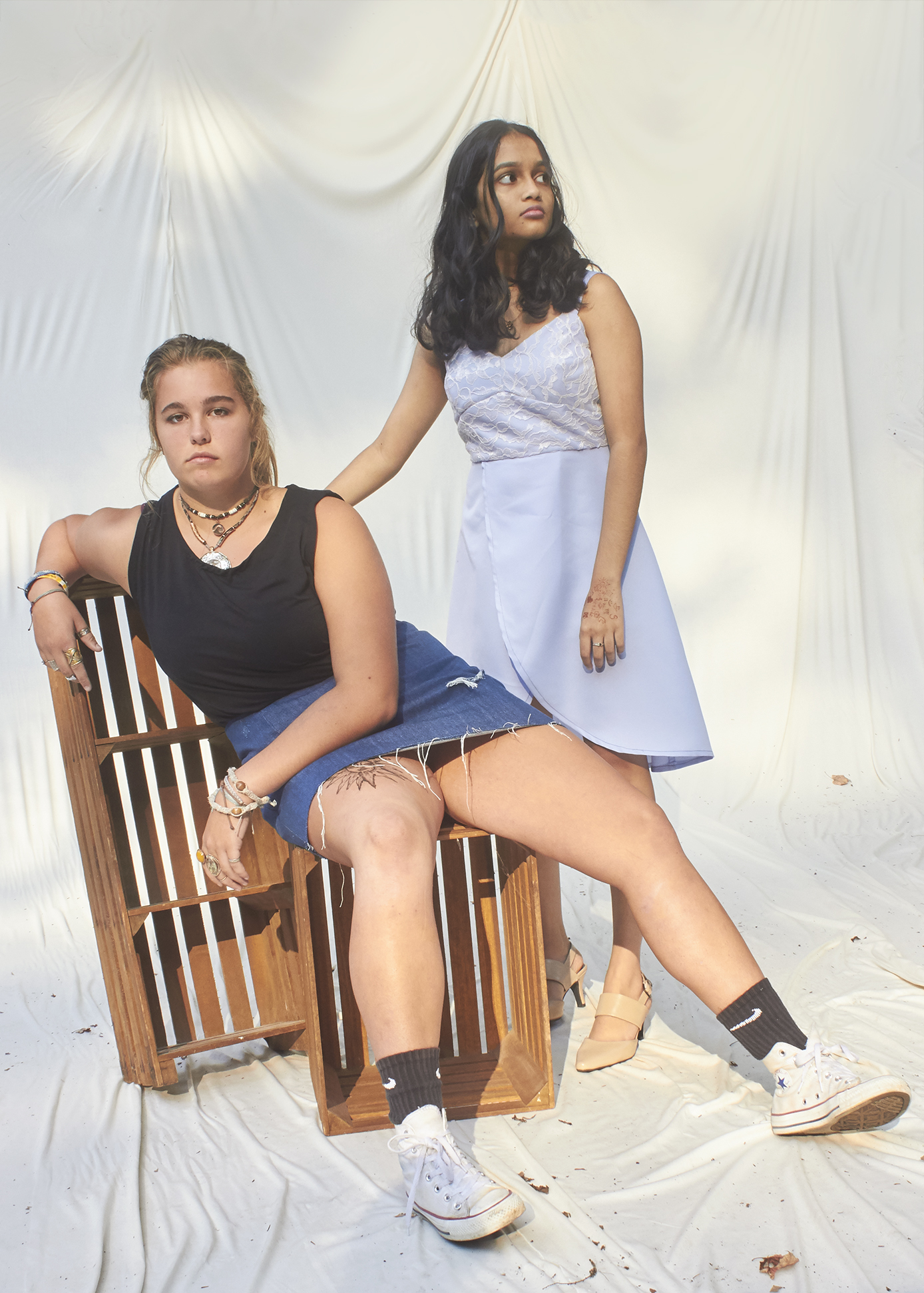 Two girls pose (one sitting on crates and the other leaning over the one seated). The seated girl is wearing a black top and distressed denim skirt while th eohter girl wears a sky blue lace overplayed dress.