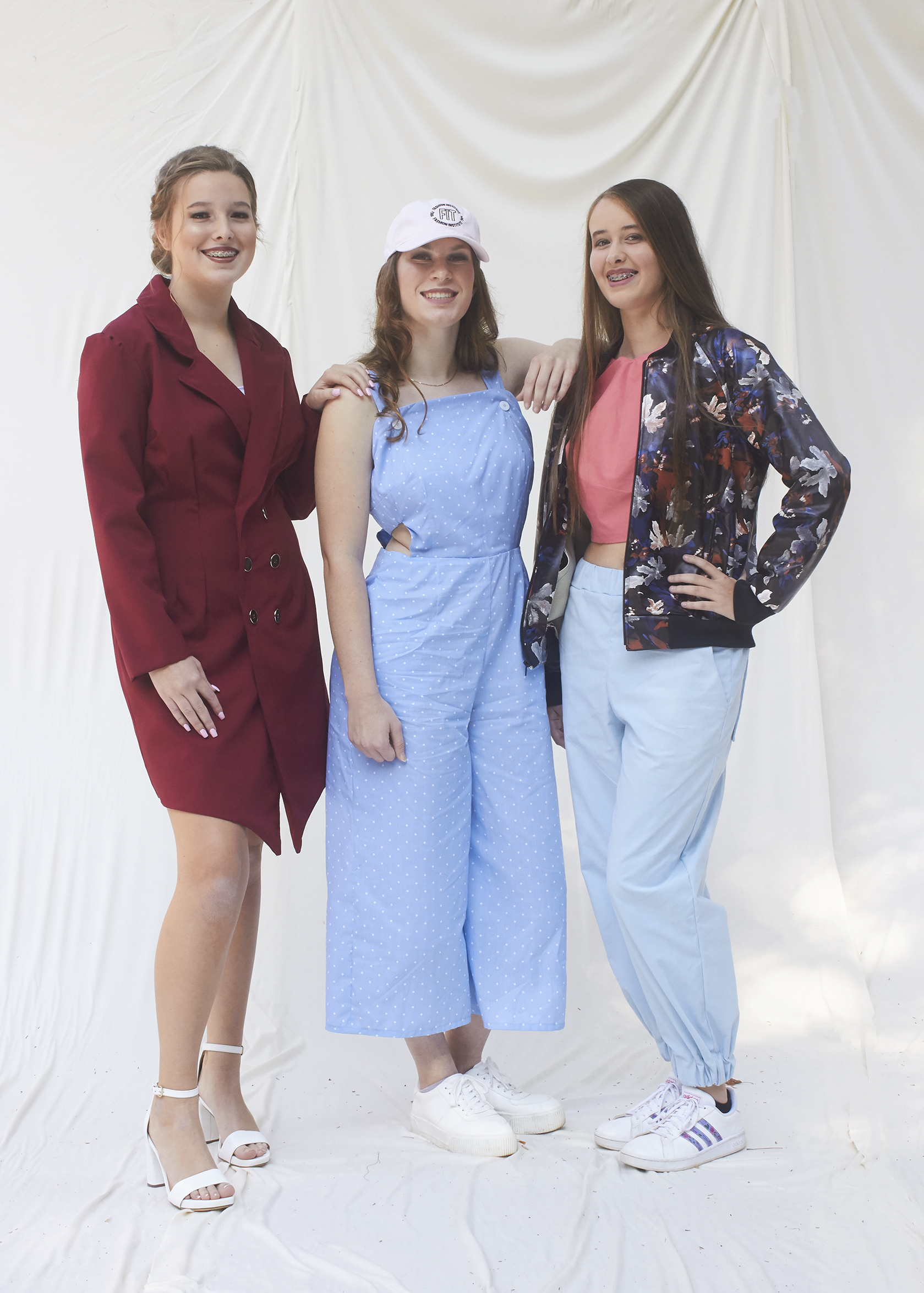 Three girls stand together in various outfits for 2020 fashion show. The left most girl wears a business suit dress, the middle girl wears a light blue jumpsuit, and the far right girl wears a pleather floral jacket and blue pants.