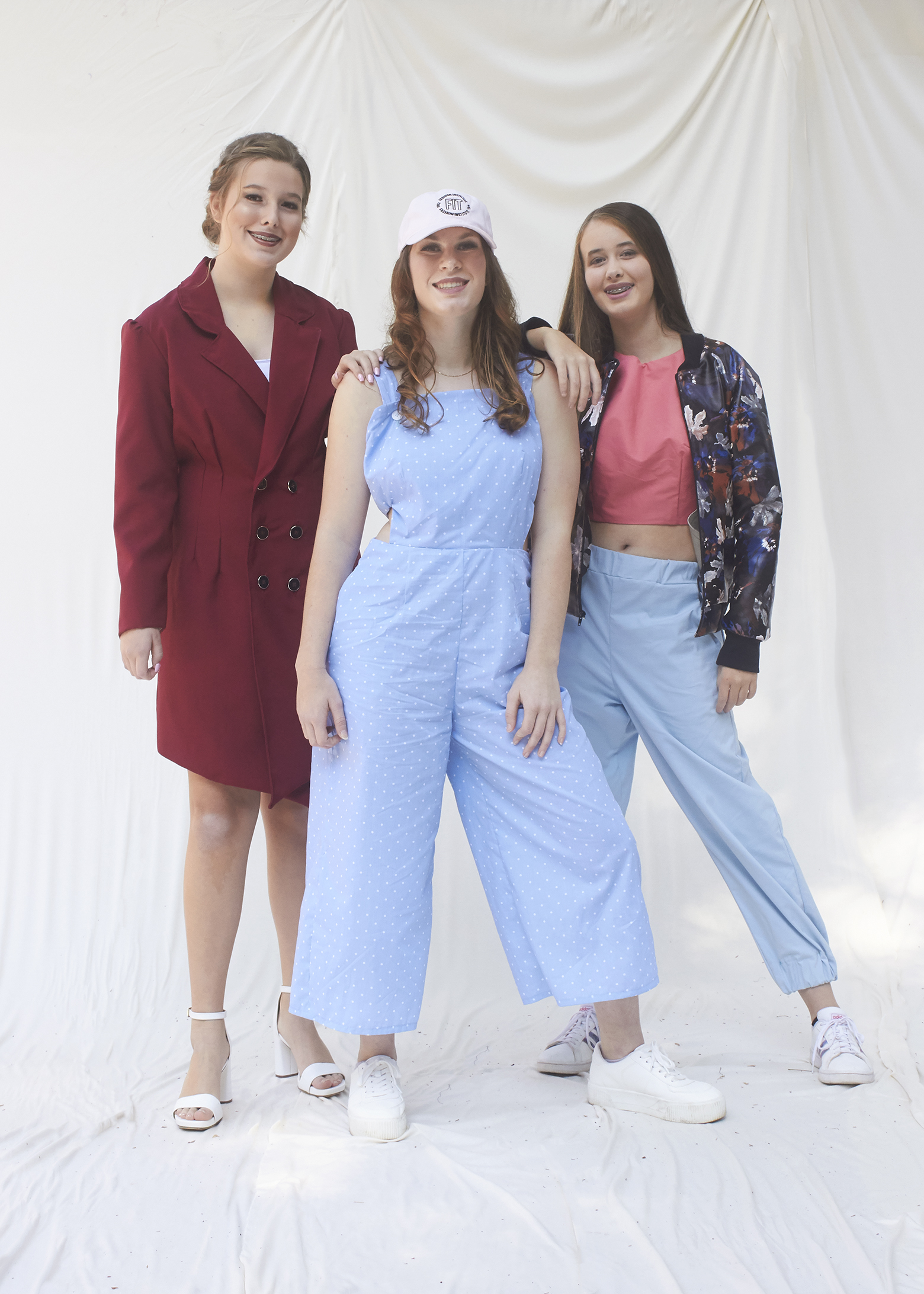 Three girls stand together in various outfits. The left most girl wears a business suit dress, the middle girl wears a light blue jumpsuit, and the far right girl wears a pleather floral jacket and blue pants.