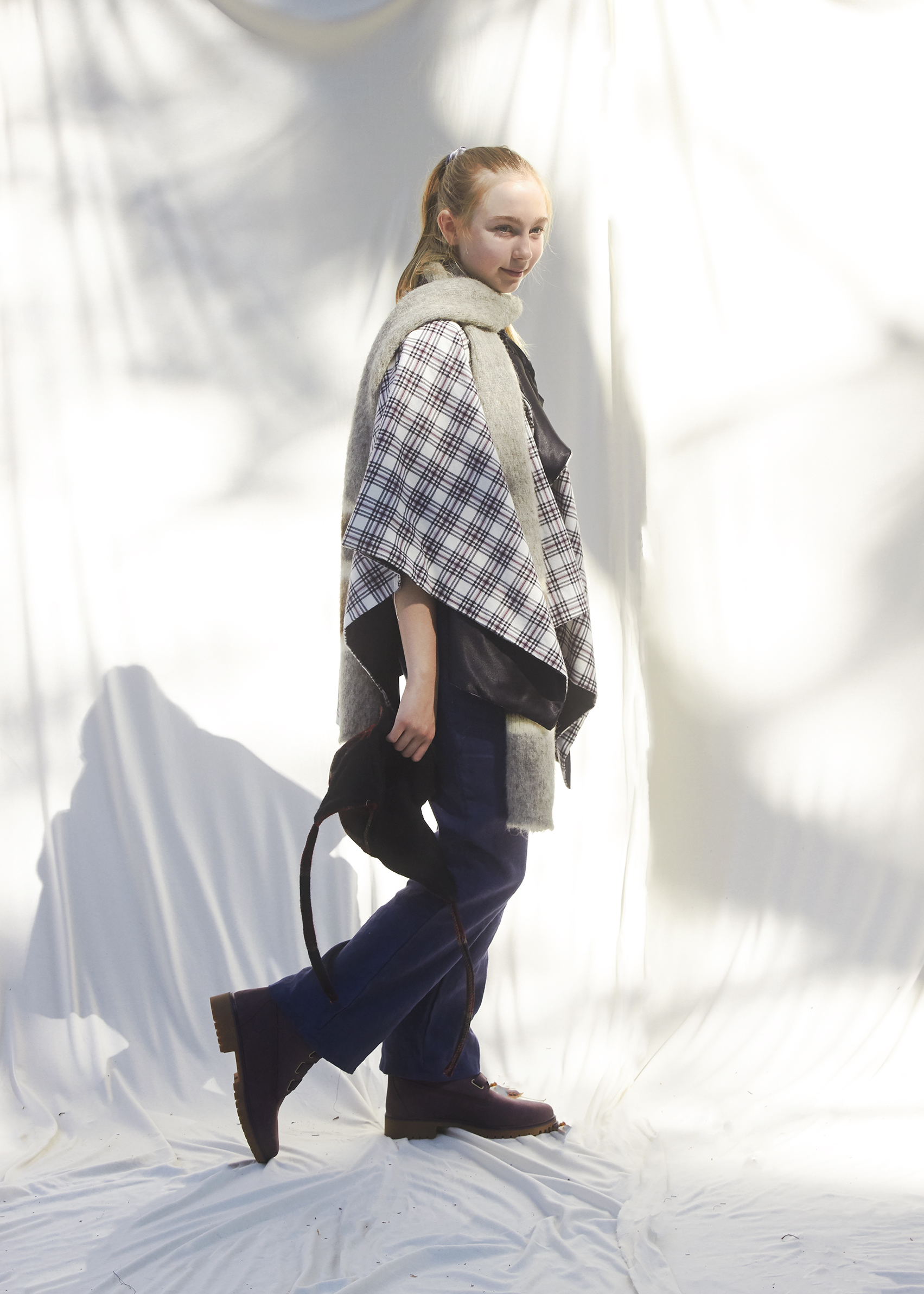 Young girl stands in winter plaid poncho and holds a plaid trapper hat in her hand