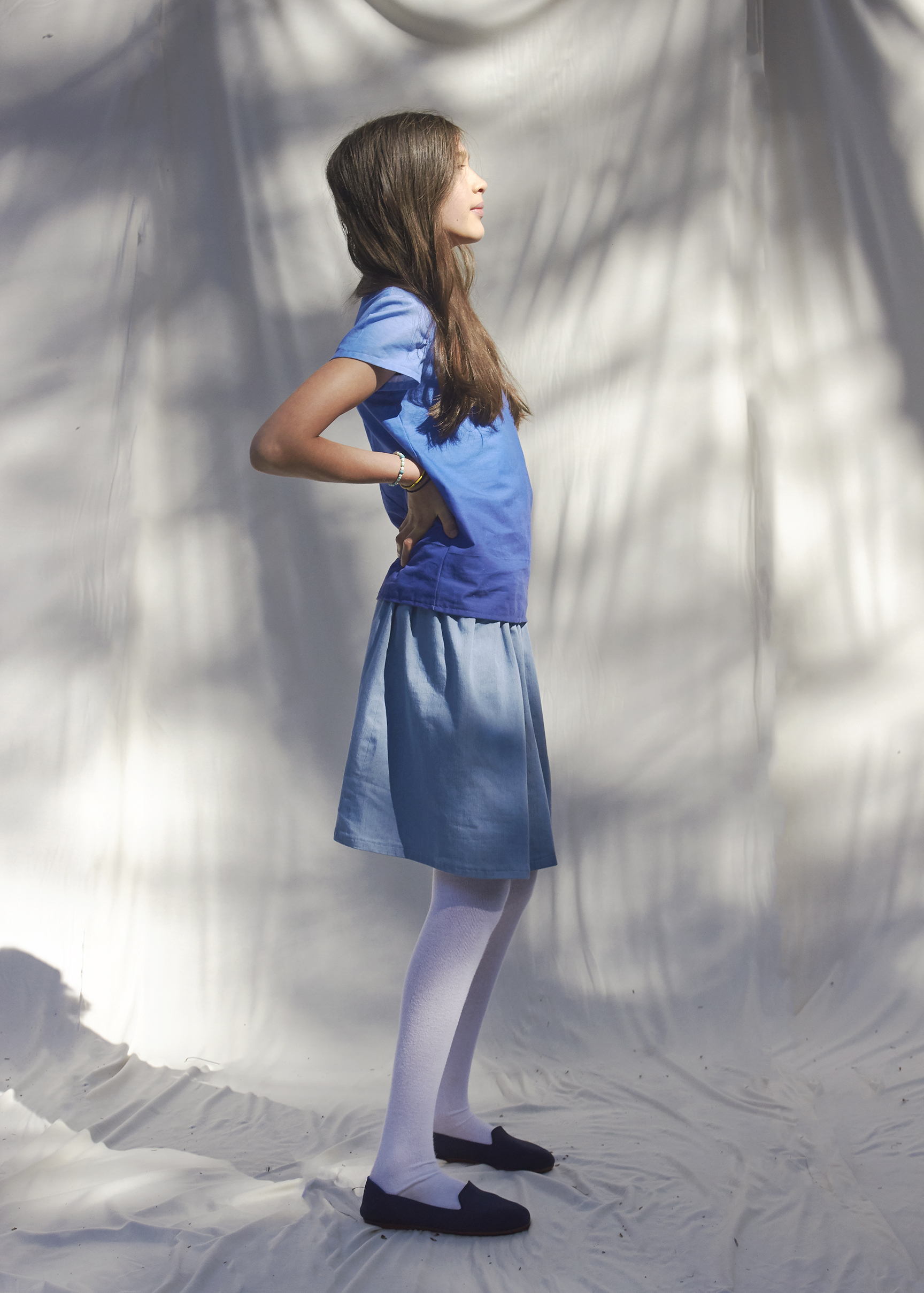 Young girl stands in profile view with hand on her back while she wears a ombre blue top and a sky blue gathered skirt