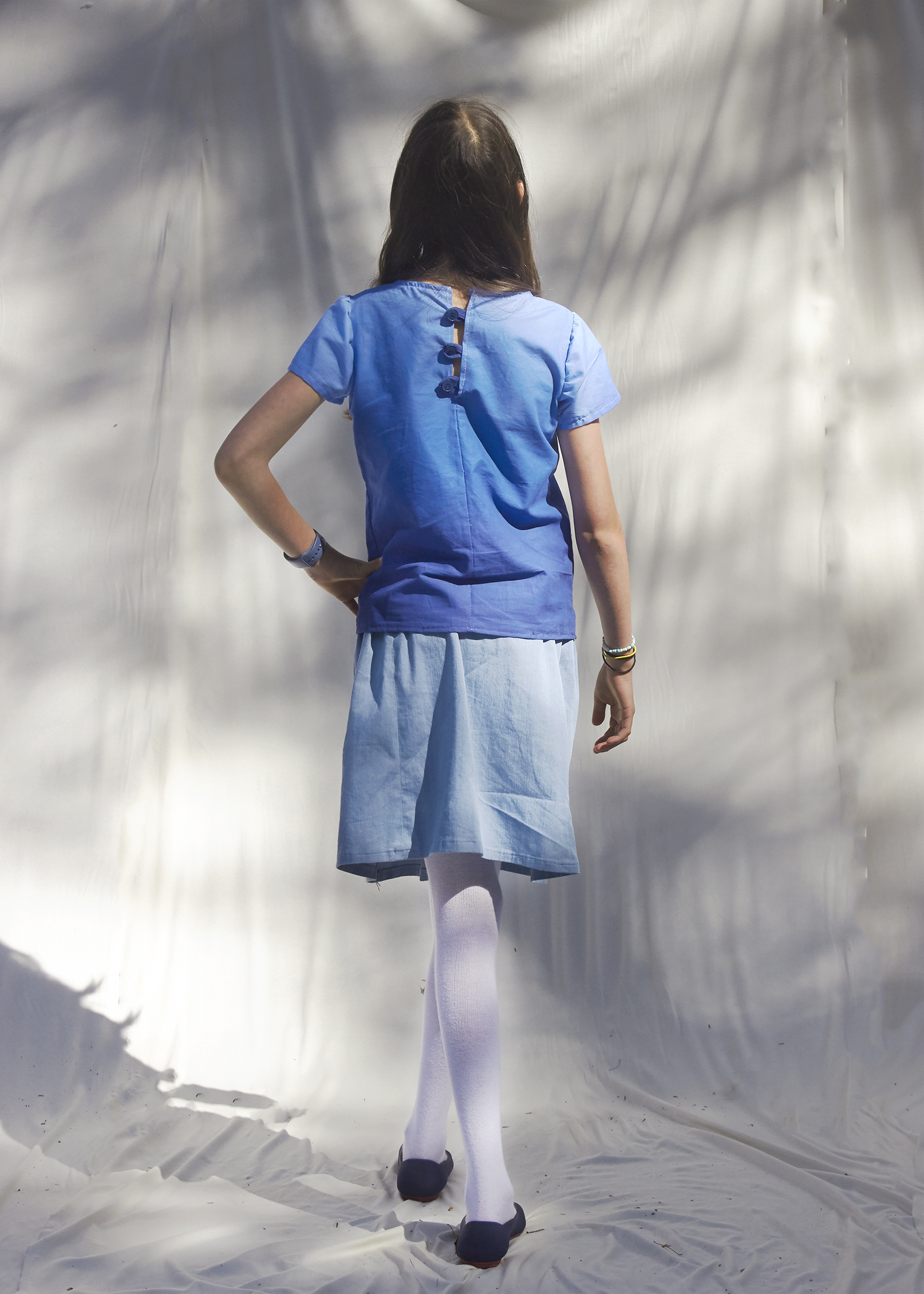 Back view of a young girl wearing a buttoned back ombre blue short sleeve shirt and sky blue gathered skirt