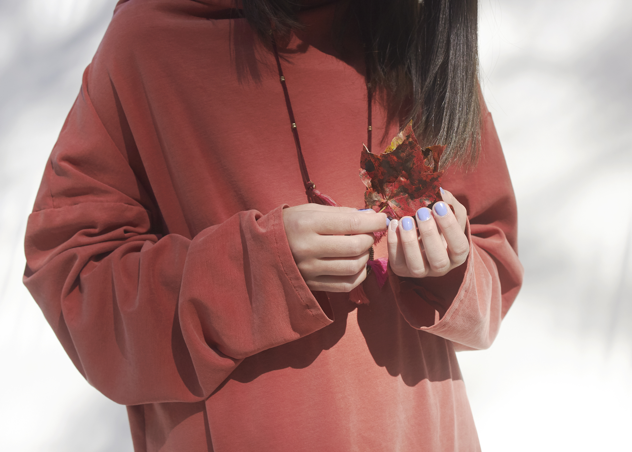 Close up photo of a girl, wearing a long sleeve burnt orange sweater dress, is holding a leaf in her hands